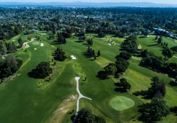 Aerial view of a sprawling golf course with green fairways and trees, surrounded by a distant cityscape